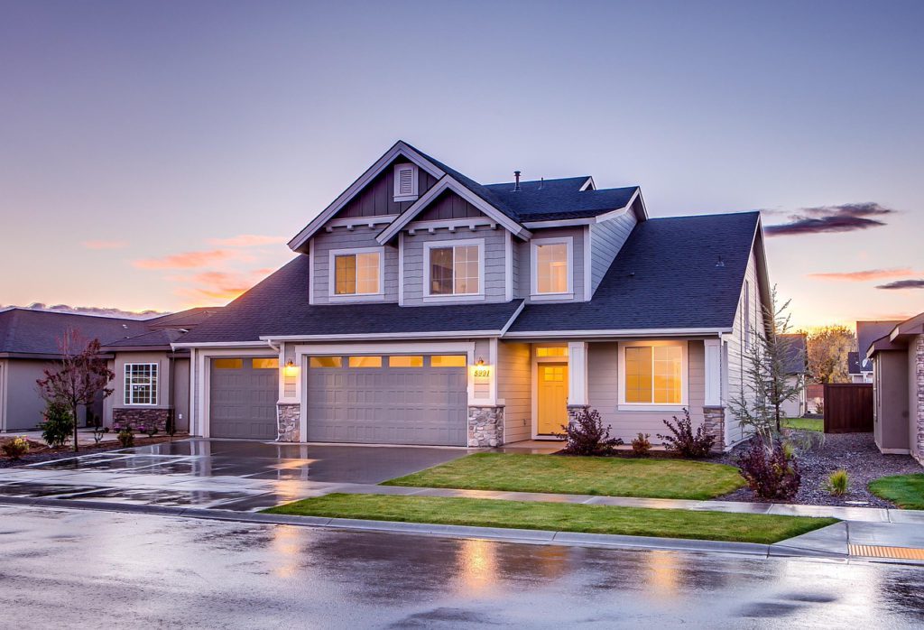 Modern two-storey suburban house with grey cladding, double garage, lit windows at dusk, and a neatly landscaped front lawn after rain